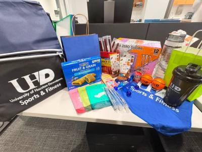 Assortment of food items, Sports & Fitness swag, and prizes. Snacks, a drawstring backpack, pens, a water bottle, a UHD Sports and Fitness drawstring backpack, and face masks on a table. The items are prizes for a gym membership drive for employees at the university of houston-downtown.