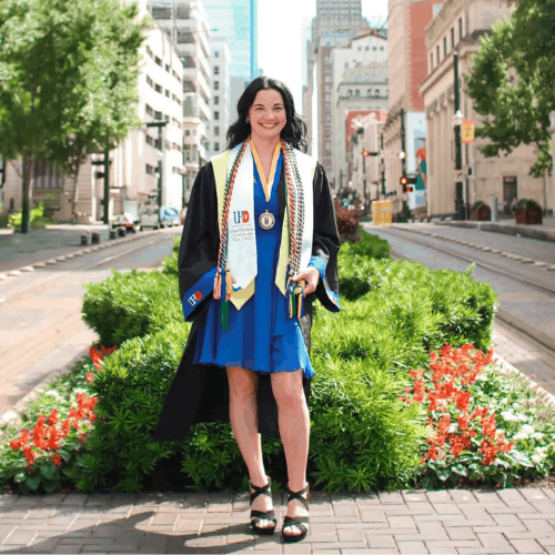 Social Work grad Amber Perez wears cords, stole, and graduation gown with University of Houston Downtown logo while smiling on Main Street in downtown Houston