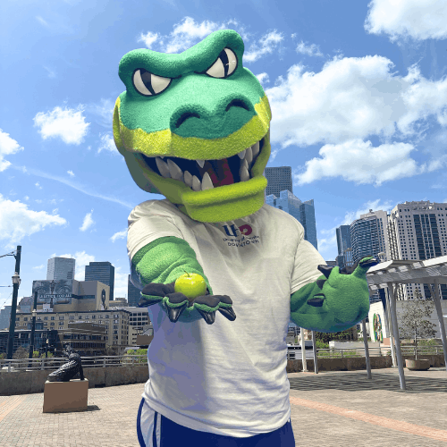 University of Houston-Downtown mascot Ed-U-Gator holds a green apple on the university's south deck with the Downtown Houston skyline behind them. 