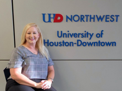 Jamie Thomas seated next to wall which reads UHD Northwest University of Houston-Downtown. Thomas is the executive director of Off-Campus Learning & Academic Partnerships for the University of Houston-Downtown.